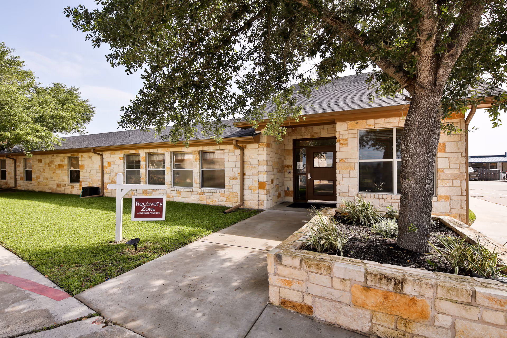 Exterior view of a single-story stone building with a sidewalk leading to a glass door entrance. There is a large tree planted in a stone-bordered garden bed near the entrance and a sign on the grass that reads 'Recovery Zone Patients At Work'.