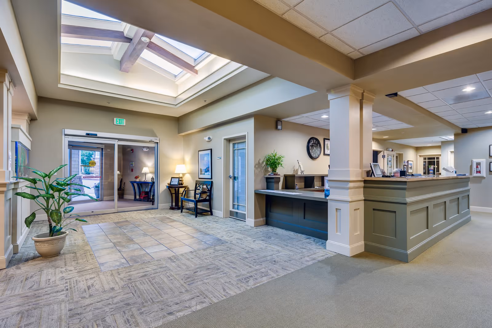 Bright and spacious reception area of Woodland Terrace with a large skylight, a tiled entrance floor, a potted plant, seating area with chairs and a lamp, and a long reception desk with decorative columns and plants.