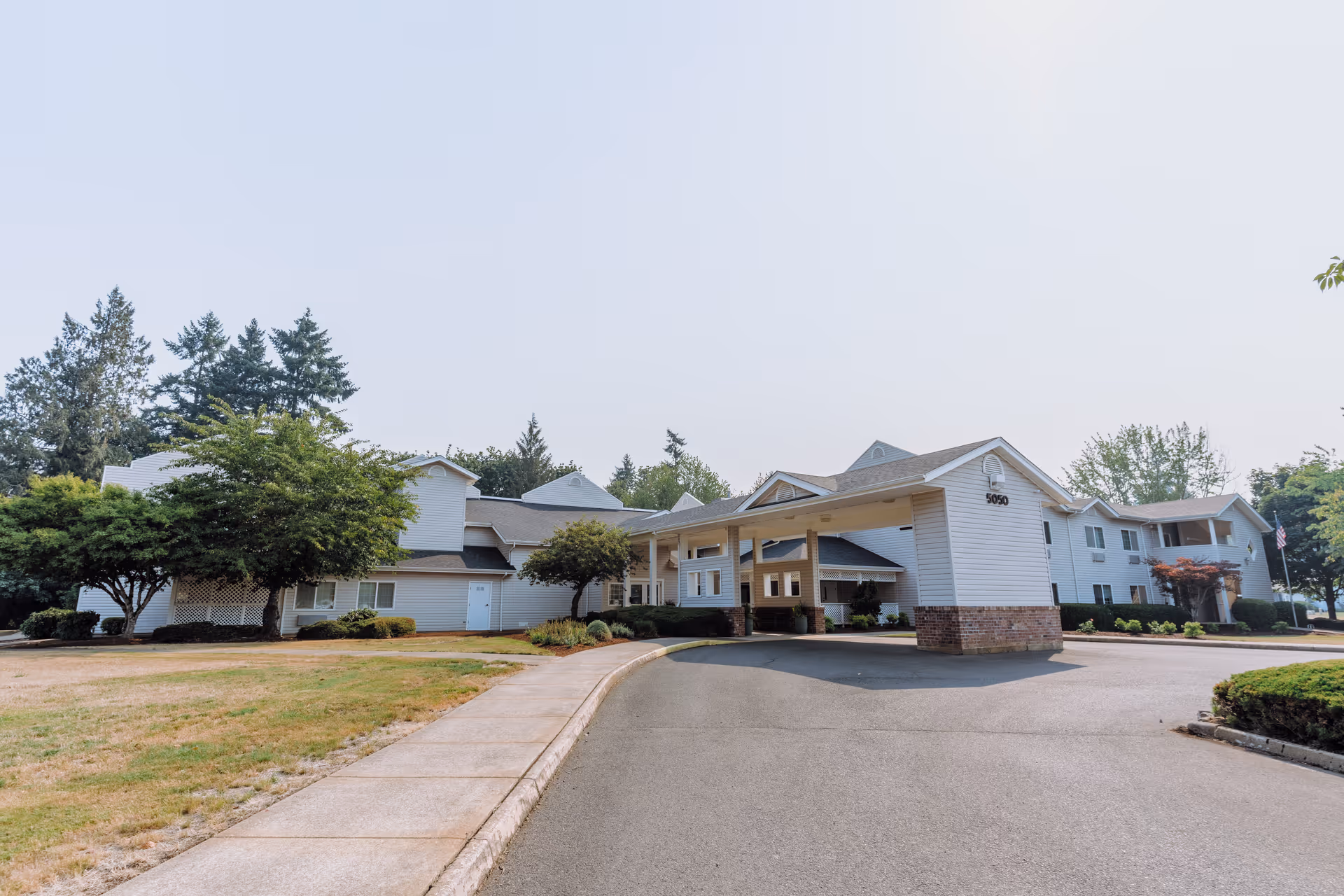 Exterior view of a senior living facility building with white siding and a covered entrance. There are trees and shrubs around the building, a paved driveway, and a sidewalk leading to the entrance.