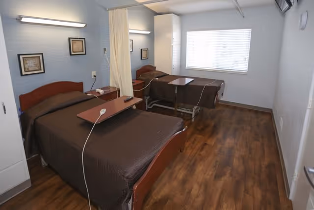 A healthcare facility room with two hospital beds separated by a curtain. Each bed has a brown blanket and an overbed table. The room has wood flooring, light blue walls, a large window with blinds, and framed pictures on the wall.