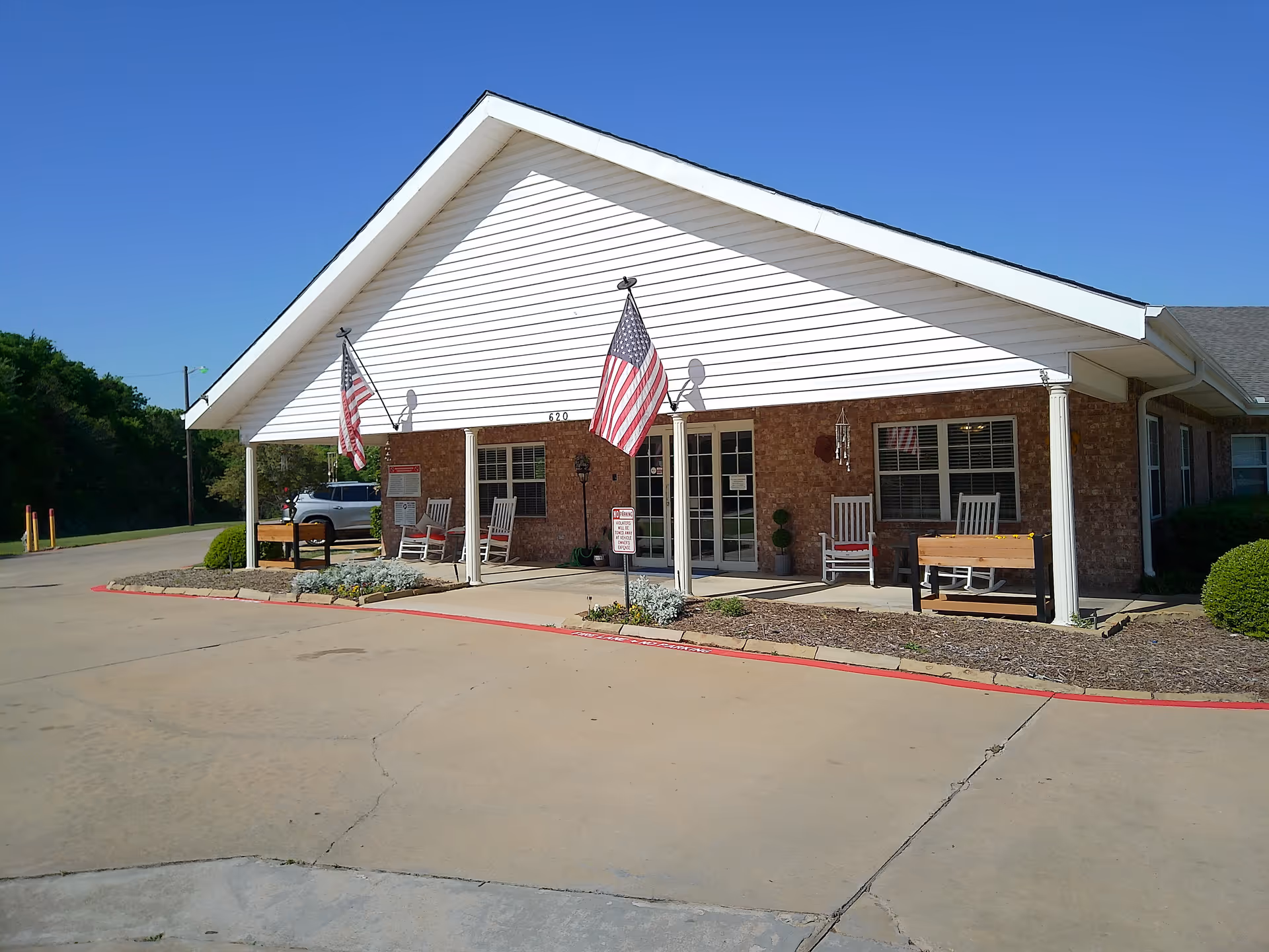 Exterior view of a single-story brick building with a white gabled roof and a covered porch. The porch has several white rocking chairs and two American flags mounted on poles. There is a small garden bed with plants along the front of the porch, and a paved driveway in front. The sky is clear and blue.