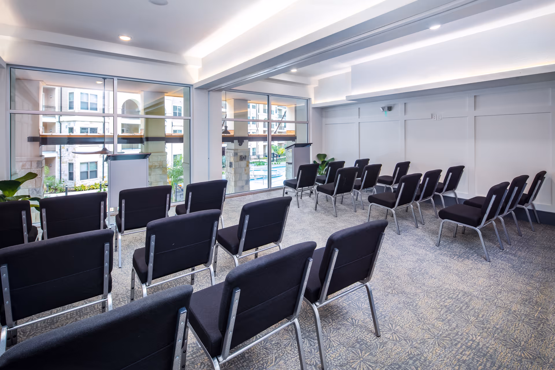 Small meeting room with rows of black chairs facing two lecterns and large windows overlooking a courtyard.