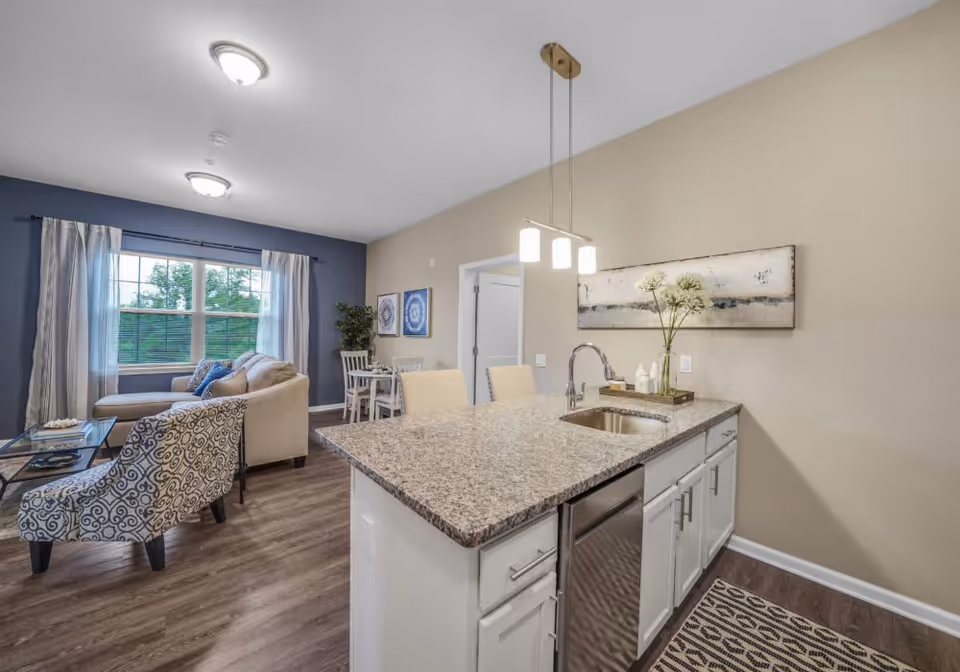 Modern open-concept living space featuring a kitchen island with granite countertop and sink, two beige chairs, and pendant lighting. Adjacent to the kitchen is a living room area with a patterned armchair, beige sofa, glass coffee table, and large window with curtains letting in natural light. The walls are painted beige and blue, with framed artwork and a potted plant in the corner.
