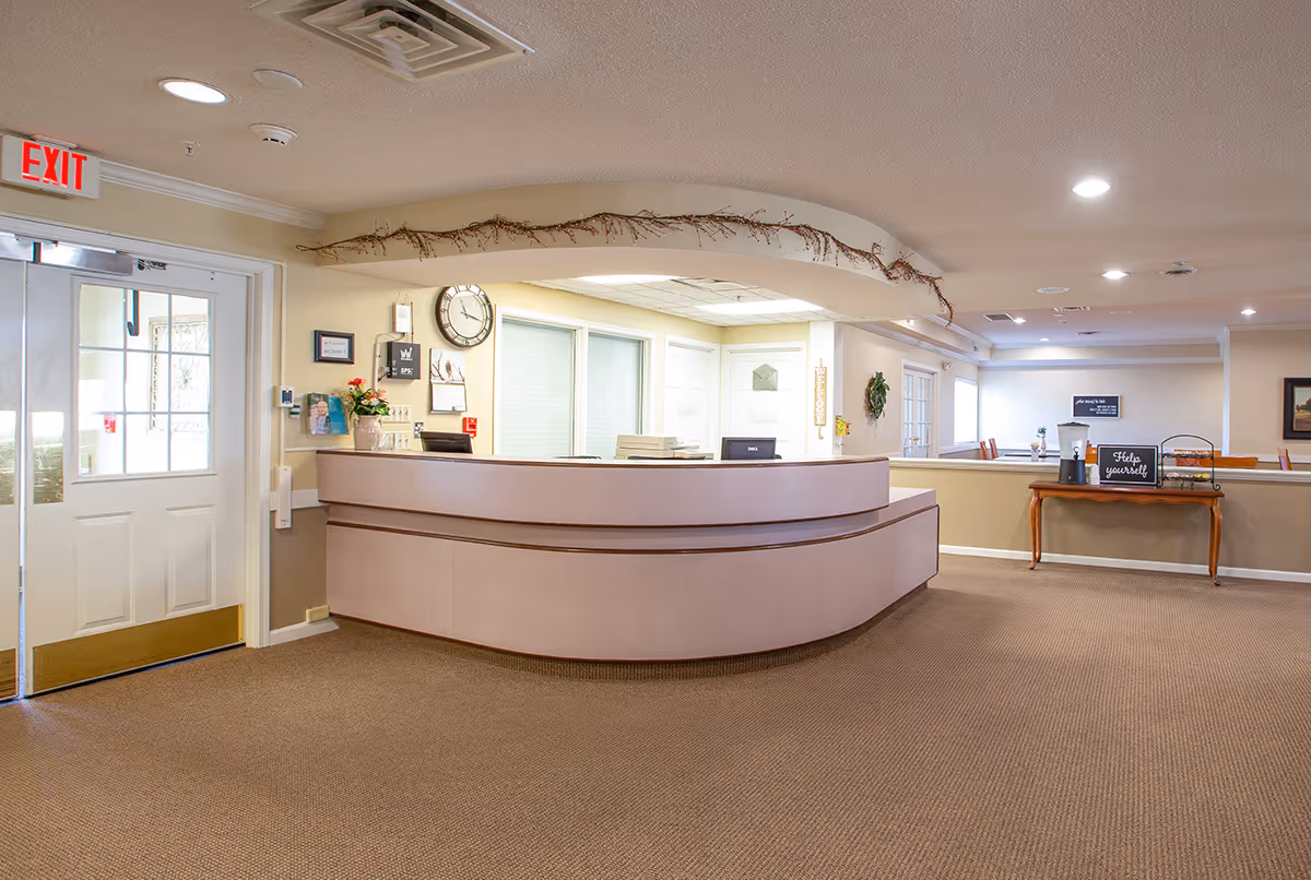 Reception area inside Pathfinder Senior Living facility with a curved front desk, a clock on the wall, a door with an exit sign above it, and a small table with a water dispenser and a sign that says 'Help yourself'.