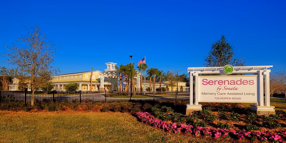 Exterior view of Serenades at West Orange, a memory care assisted living facility, showing a large building with a yellow facade, palm trees, an American flag, and a white sign with the facility's name and address surrounded by landscaped flowers and greenery under a clear blue sky.