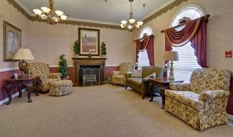 Common room with floral upholstered armchairs and a sofa arranged around a decorative fireplace beneath arched windows with drapes and chandeliers.