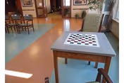 Interior view of a senior living facility common area with a checkers game table in the foreground, chairs around it, and additional seating and tables in the background. The floor has a colorful pattern with blue and orange sections, and there are plants and framed pictures on the walls.