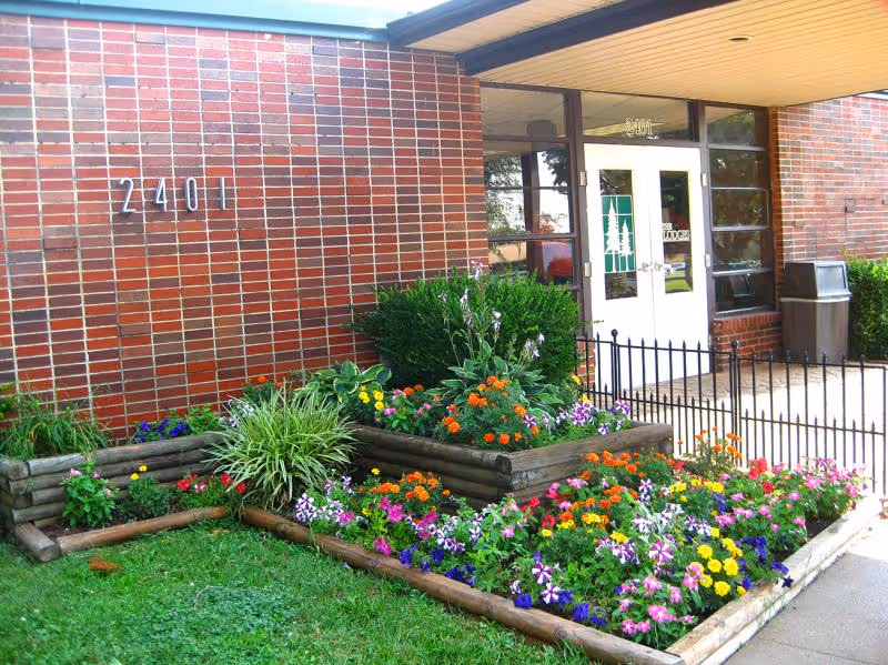 Entrance of a brick building numbered 2401 with double doors and colorful flower beds in front.
