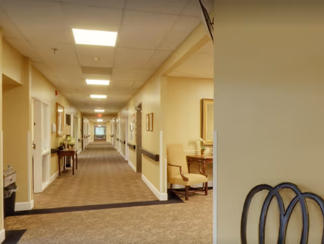 Long carpeted interior hallway of an assisted living facility with doors on both sides and a seating area visible to the right.