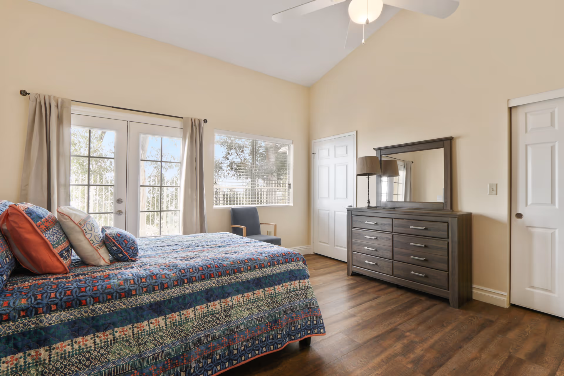 A bright bedroom with a large bed covered in a colorful patterned quilt and multiple pillows. The room has wooden flooring, a dark wooden dresser with a mirror and a lamp on top, a blue armchair near a window with blinds, and French doors with beige curtains. The walls are painted light beige and there is a ceiling fan above.