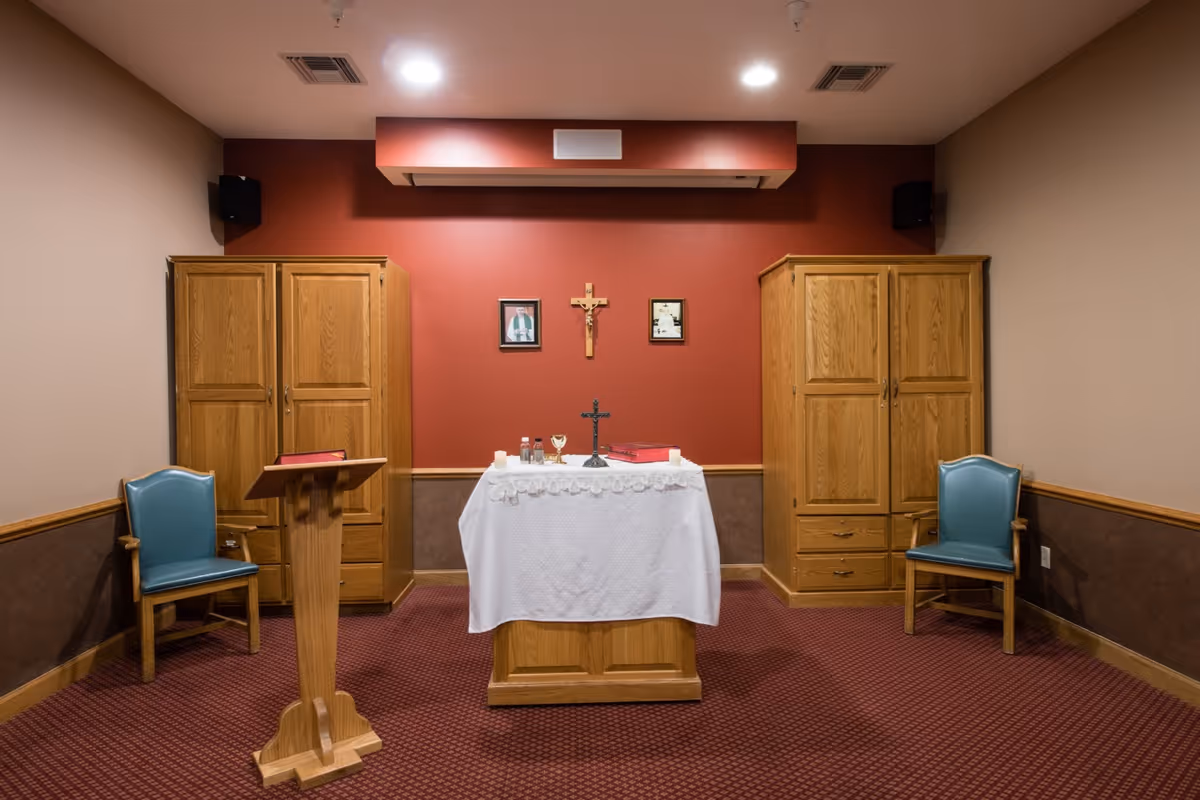 A small chapel room with a red accent wall featuring a crucifix and two framed pictures. In the center, there is an altar covered with a white cloth holding religious items including a cross, candles, and a chalice. On either side of the altar are wooden cabinets and blue cushioned chairs. A wooden lectern with a red book is positioned to the left of the altar. The room has a carpeted floor and beige walls with wood trim.