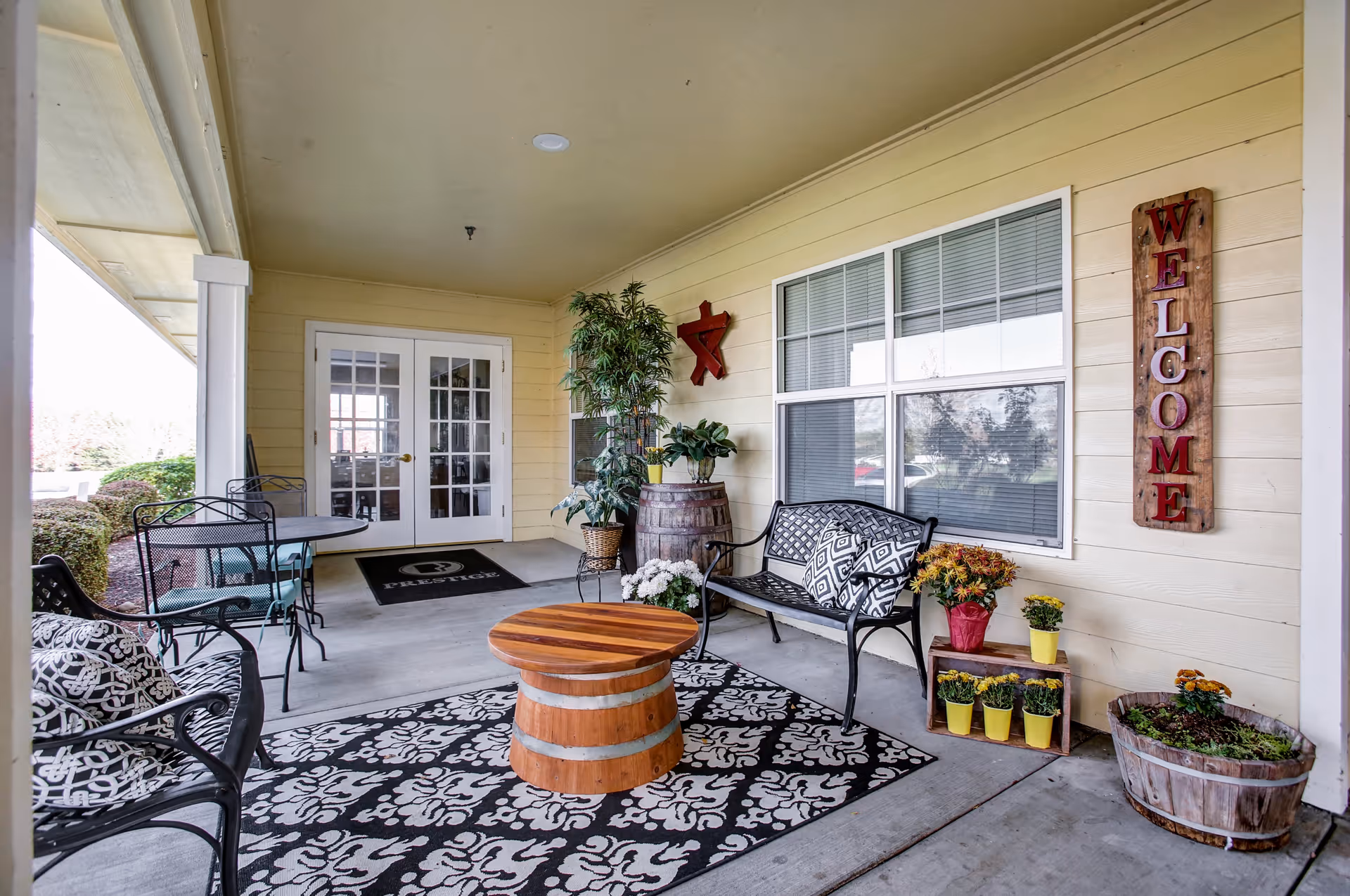 Covered outdoor seating area at a senior living facility with a black and white patterned rug, a round wooden table, black metal chairs and bench with patterned cushions, potted plants, and a wooden sign on the wall that says 'WELCOME'. Double glass doors lead inside the building.
