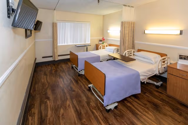 A healthcare facility room with two hospital beds, each with a purple blanket and white pillow. The room has wood-patterned flooring, a window with vertical blinds, wall-mounted lights above each bed, a mounted TV on the wall, and a privacy curtain between the beds. There is also a small wooden cabinet and a table on wheels beside one of the beds.
