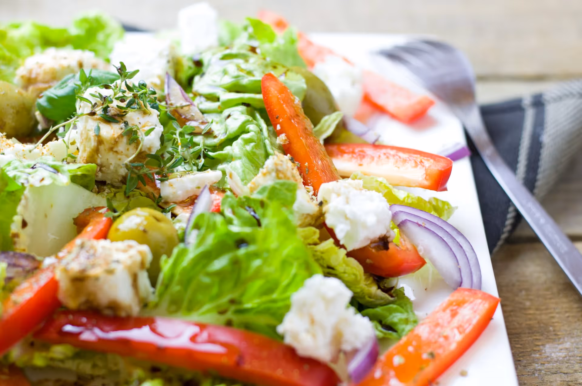 Close-up of a fresh salad on a white plate featuring lettuce, red bell pepper slices, red onion, green olives, and crumbled white cheese, with a fork and a napkin in the background on a wooden surface.