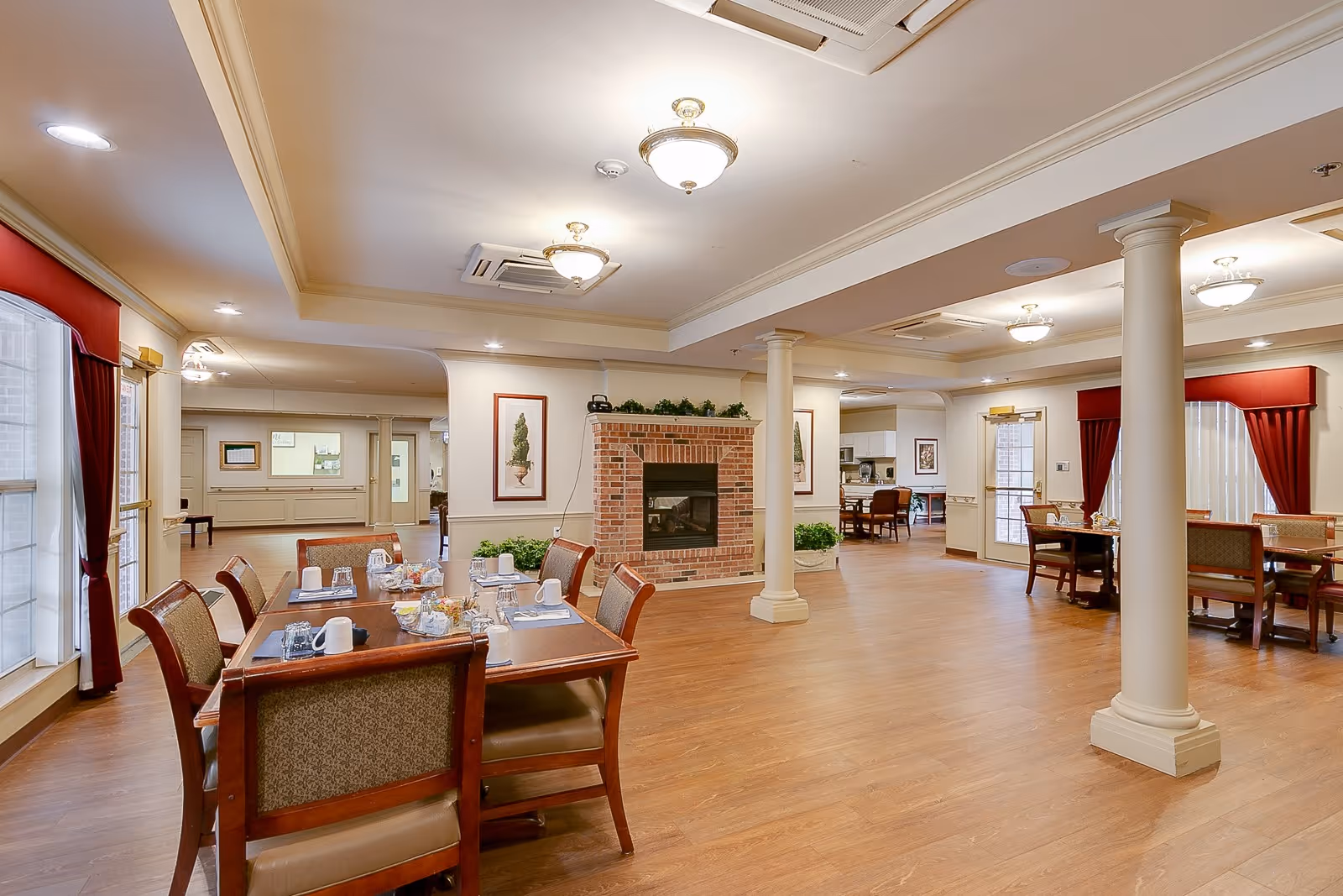 A spacious senior living dining area with wooden floors, multiple dining tables set with cups and glasses, a brick fireplace in the center, cream-colored walls, and red curtains on the windows. The room is well-lit with ceiling lights and has decorative plants and framed artwork on the walls.
