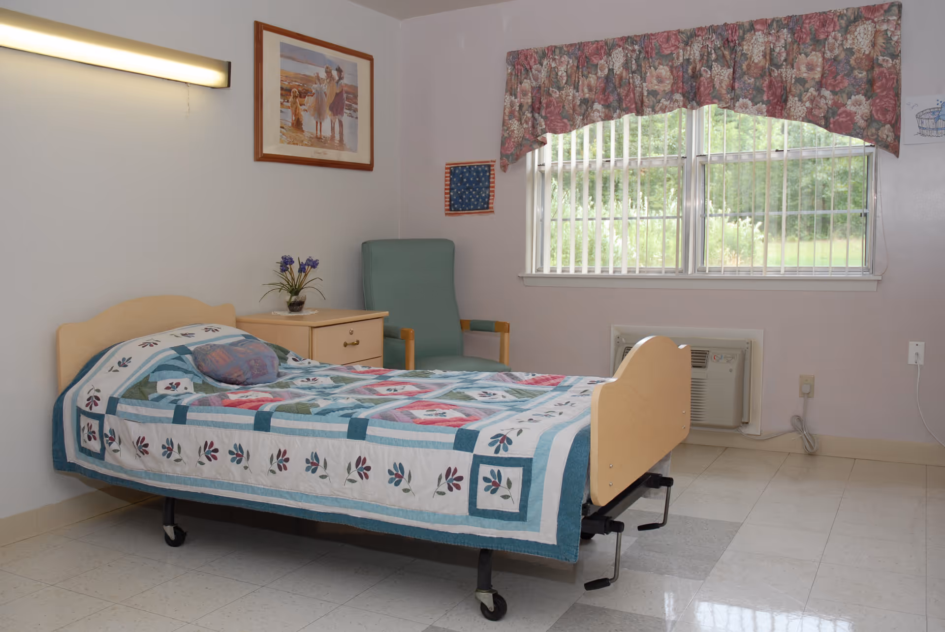 A simple bedroom in a senior living facility with a single bed covered by a floral quilt, a wooden nightstand with a small potted plant, a green armchair, a large window with vertical blinds and floral valance, and a wall-mounted air conditioning unit below the window.