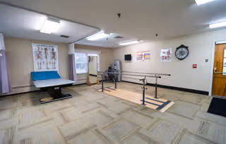 A physical therapy or rehabilitation room with parallel bars for walking exercises, an examination table with a blue cover, anatomical charts on the walls, a clock, and a door leading outside or to another room. The room has beige walls and carpeted flooring with a wooden section under the parallel bars.