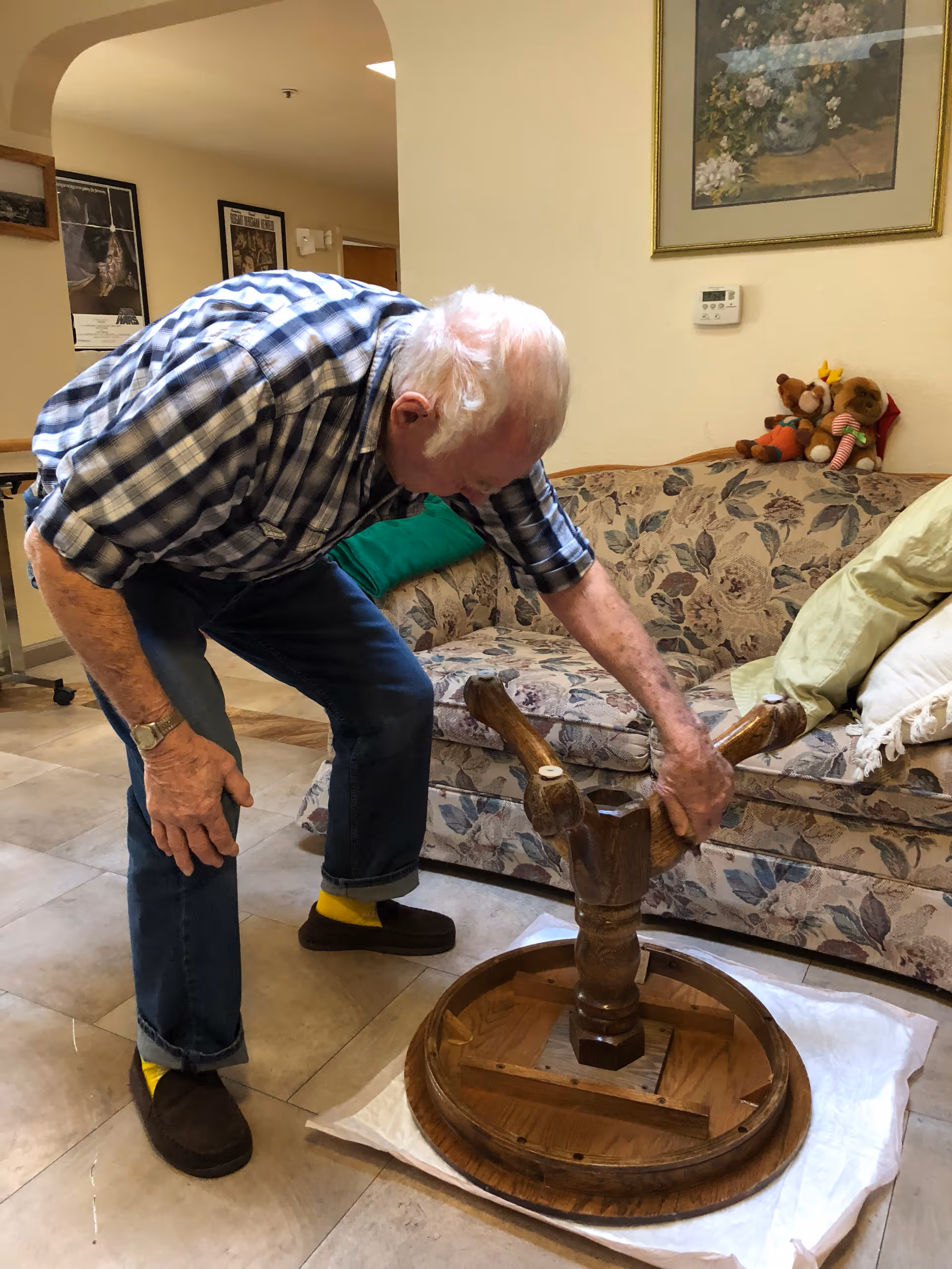 An elderly man wearing a plaid shirt, jeans, and yellow socks is bending over and working on assembling or fixing a wooden round table that is upside down on the floor. Behind him is a floral patterned couch with pillows and stuffed animals, and a framed painting on the wall.