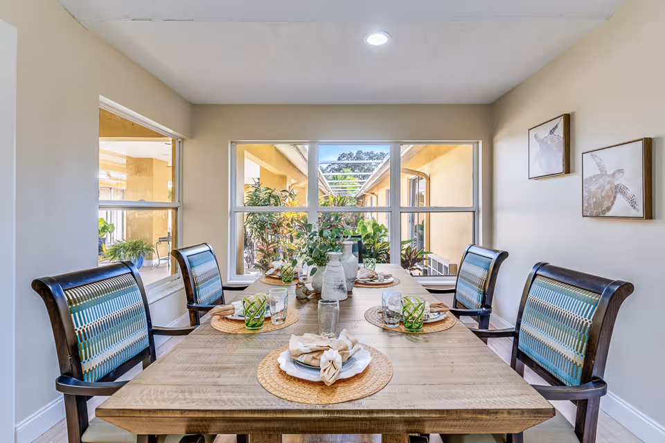 A dining room with a wooden table set for four, featuring woven placemats, plates with folded napkins, glasses, and green patterned drinking glasses. Four wooden chairs with patterned cushions surround the table. Large windows provide a view of an outdoor garden area with plants. Two framed pictures of turtles hang on the beige wall to the right.