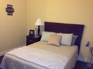 Small bedroom with a made bed, wooden headboard, nightstand with lamp and framed photos, and wall decor reading 'Hope Faith Peace'.