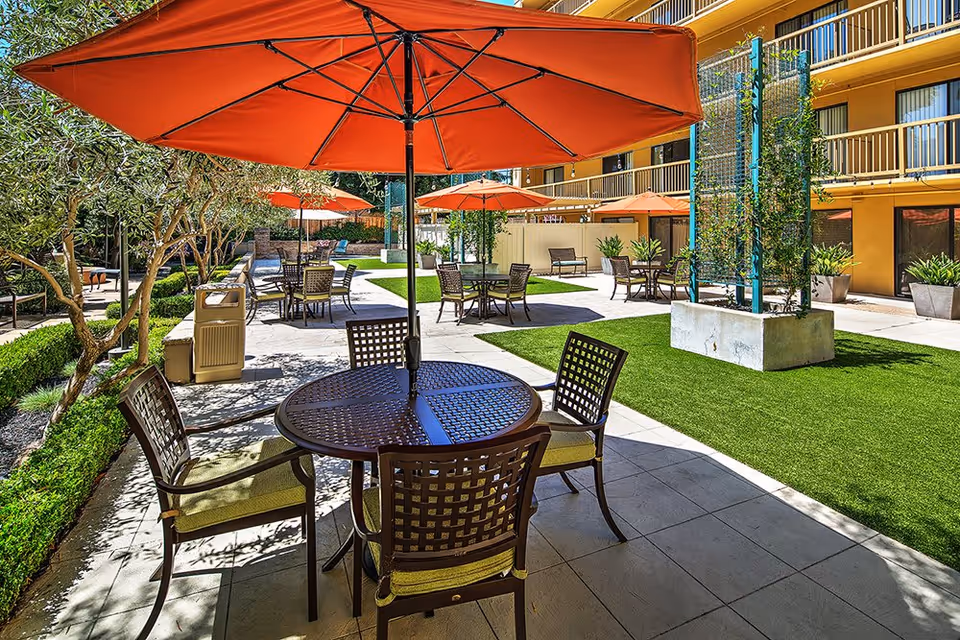 Outdoor patio area at The Point At Rockridge Senior Living with multiple round tables and chairs under large orange umbrellas, surrounded by greenery and a multi-story building in the background.