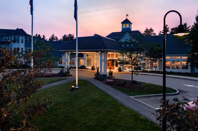 Exterior view of a senior living facility building at dusk with a covered entrance, lit windows, a street lamp, flagpoles, and landscaped greenery.