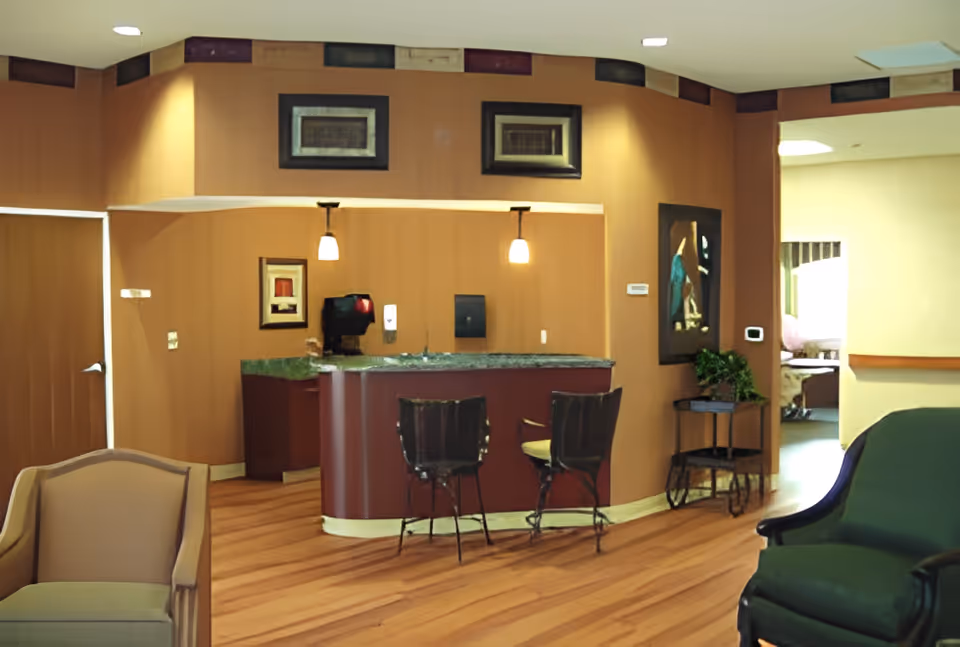 Interior view of a senior living facility lounge area with wooden flooring, a small bar counter with two chairs, framed artwork on the walls, and comfortable seating including a beige armchair and a green armchair. There is a doorway leading to another room in the background.