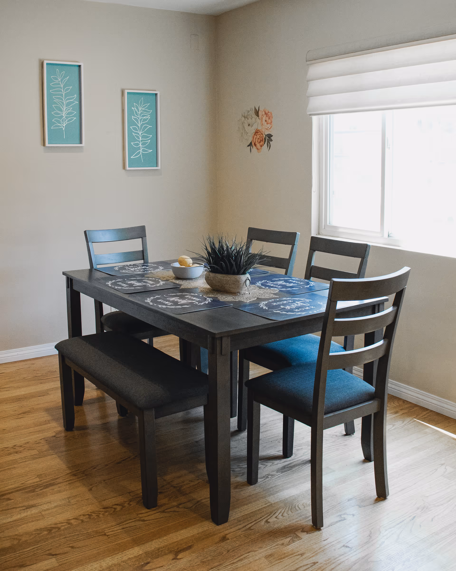 A dining area with a dark wooden table set with four chairs and a bench. The table has placemats and a centerpiece with a potted plant and a bowl with lemons. The room has light-colored walls with two framed botanical prints and a floral wall decal near a window with a white blind. The floor is wooden.