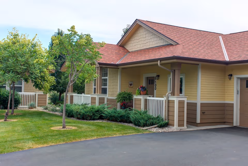 Exterior view of a single-story senior living facility building with beige and brown siding, a red shingled roof, a small porch with white railing, and green landscaping including trees and bushes.