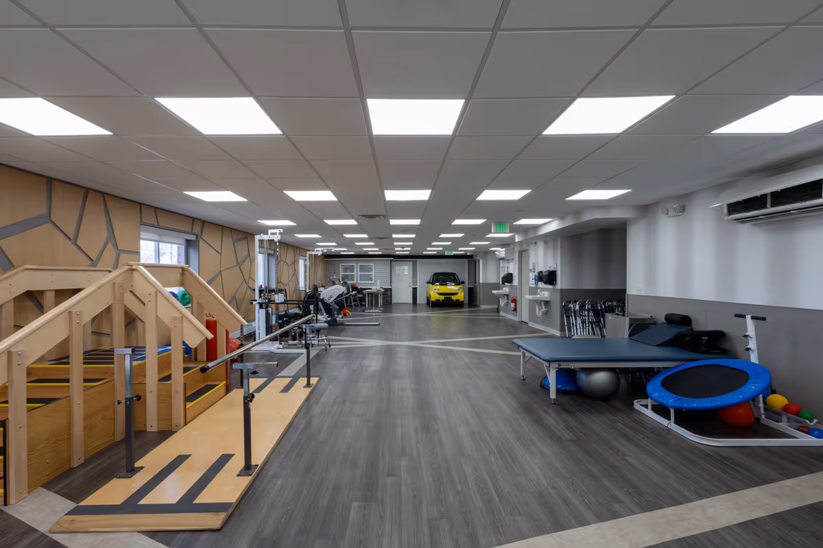 An expansive indoor rehabilitation gym with parallel bars, therapy equipment, and a bright yellow car model visible at the far end.