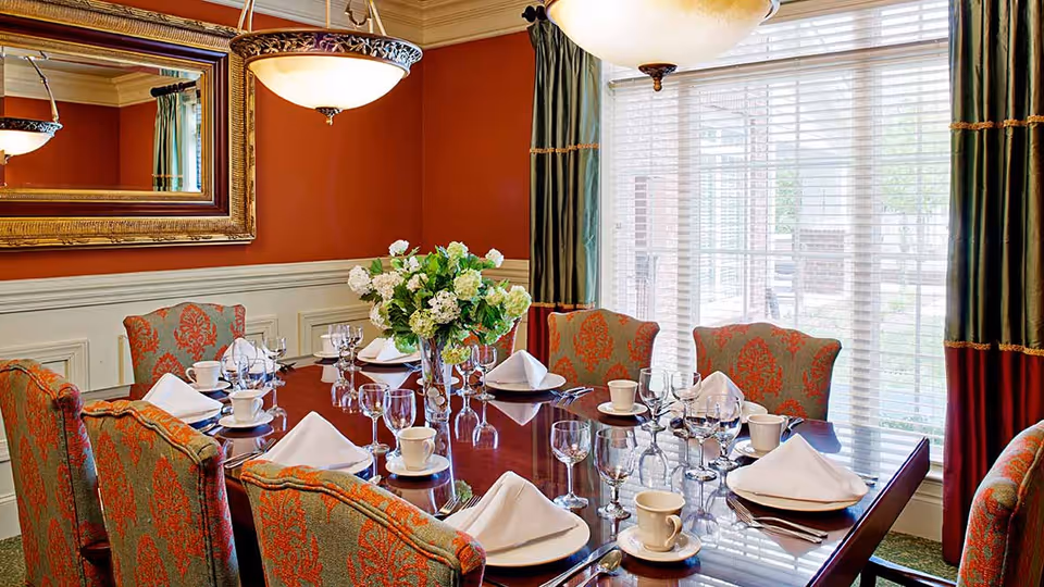 Elegant dining room with a polished wooden table set for eight, featuring white napkins, cups, saucers, and glassware. The room has red and green patterned upholstered chairs, a large floral centerpiece, a large mirror on the red wall, and large windows with green and red curtains allowing natural light to enter.