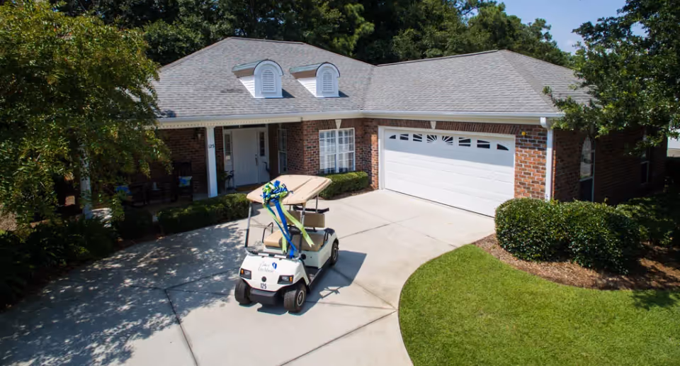 A brick building with a sloped roof and a front porch, featuring a golf cart parked in the driveway surrounded by greenery.