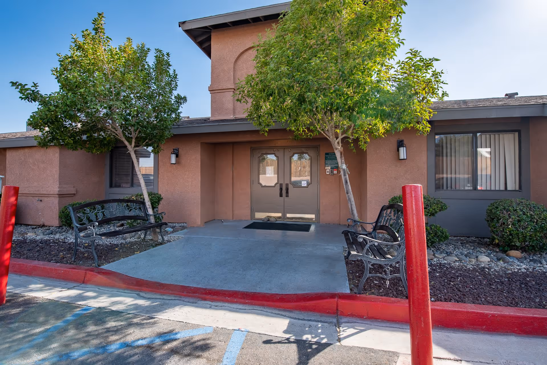 Exterior entrance of a building with double glass doors, flanked by two trees and two black metal benches on either side of the walkway. The building has a brown stucco finish and a window with vertical blinds to the right. There are red painted curbs and a parking area in front.