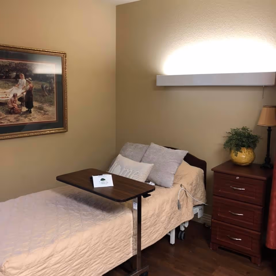 A small adjustable patient bedroom with a single bed, overbed table, pillows, a framed painting on the wall, and a nightstand with a lamp and potted plant.