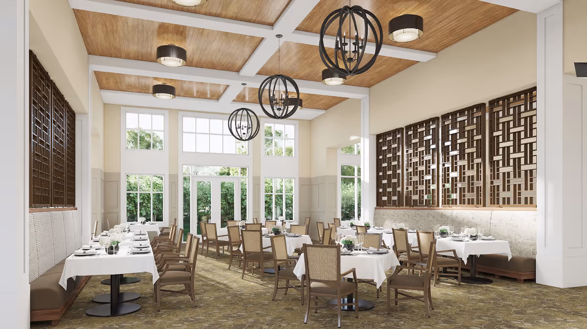 A bright and spacious dining room with multiple tables covered in white tablecloths, set with plates, glasses, and cutlery. The room features large windows allowing natural light to fill the space, wooden ceiling panels with modern circular chandeliers, and decorative wooden lattice panels on one wall. Chairs are arranged neatly around the tables, and the carpet has a patterned design.
