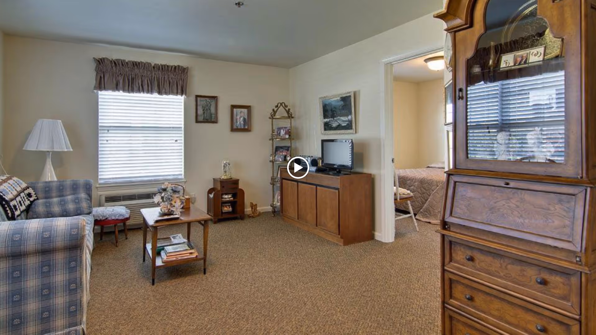 A cozy living room in a senior living facility with a blue plaid sofa, a wooden coffee table with books and a floral arrangement, a floor lamp, and a window with blinds and a valance. There is a wooden cabinet with a small TV and decorative items, a metal shelf with framed photos, and a wooden secretary desk with drawers. An open doorway leads to a bedroom with a bed and a chair.