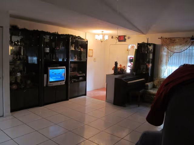 A dimly lit living room area with white tiled floor, dark wood cabinets with glass doors displaying various items, a small television, a piano with a stool, a floral couch near a window with blinds and a sheer curtain, and an exit door with an illuminated exit sign above it.
