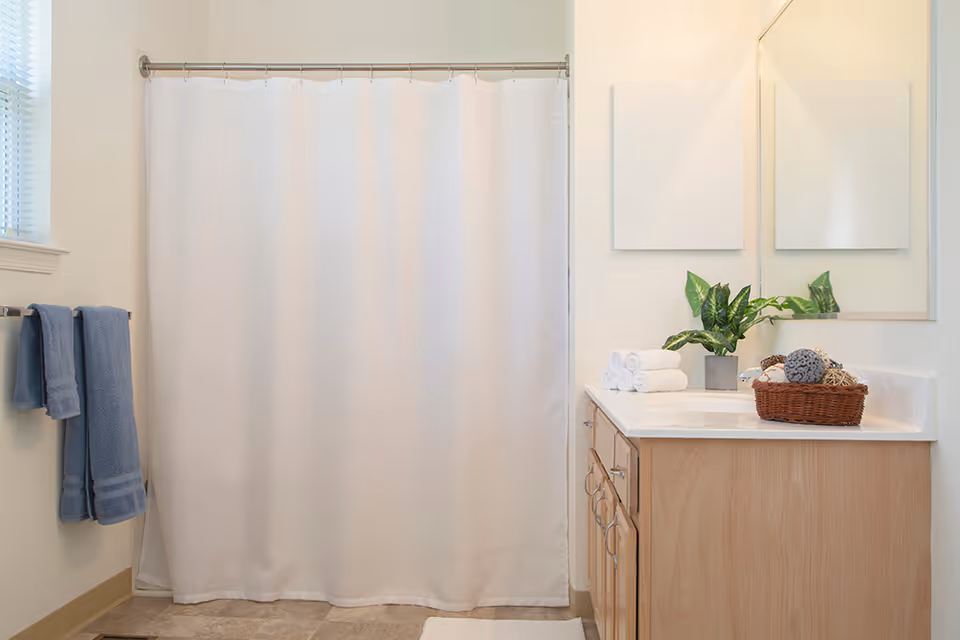 Bright bathroom with a white shower curtain, wooden vanity with sink and mirror, folded towels and a potted plant on the counter.