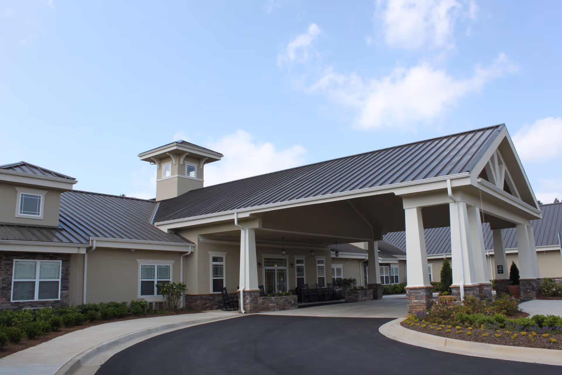 Exterior view of Benton House of Newnan, showing the building's entrance with a covered driveway, stone and beige siding, multiple windows, and a clear blue sky with some clouds.