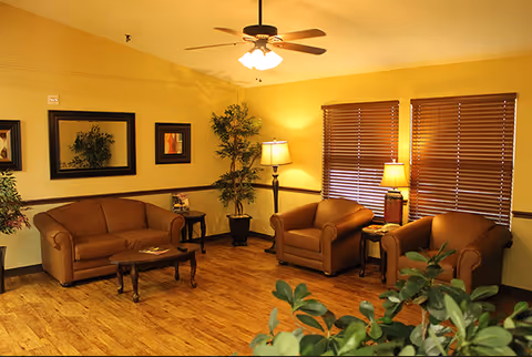 A cozy living room with a brown sofa, two matching armchairs, a wooden coffee table, side tables with lamps, a ceiling fan with light, framed artwork on the walls, and potted plants. The room has wooden flooring and windows with closed wooden blinds.