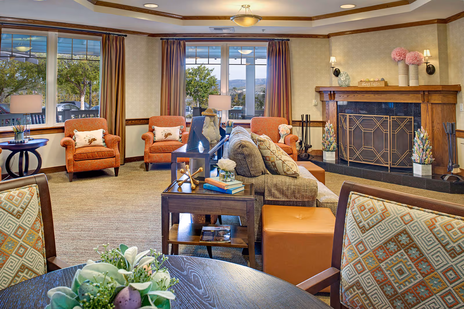 A cozy living room in a senior living facility with large windows showing trees outside. The room features a fireplace with decorative items on the mantel, three orange armchairs with patterned pillows, a gray sofa with patterned cushions, a wooden side table with books and decor, and a round table with a floral centerpiece in the foreground.