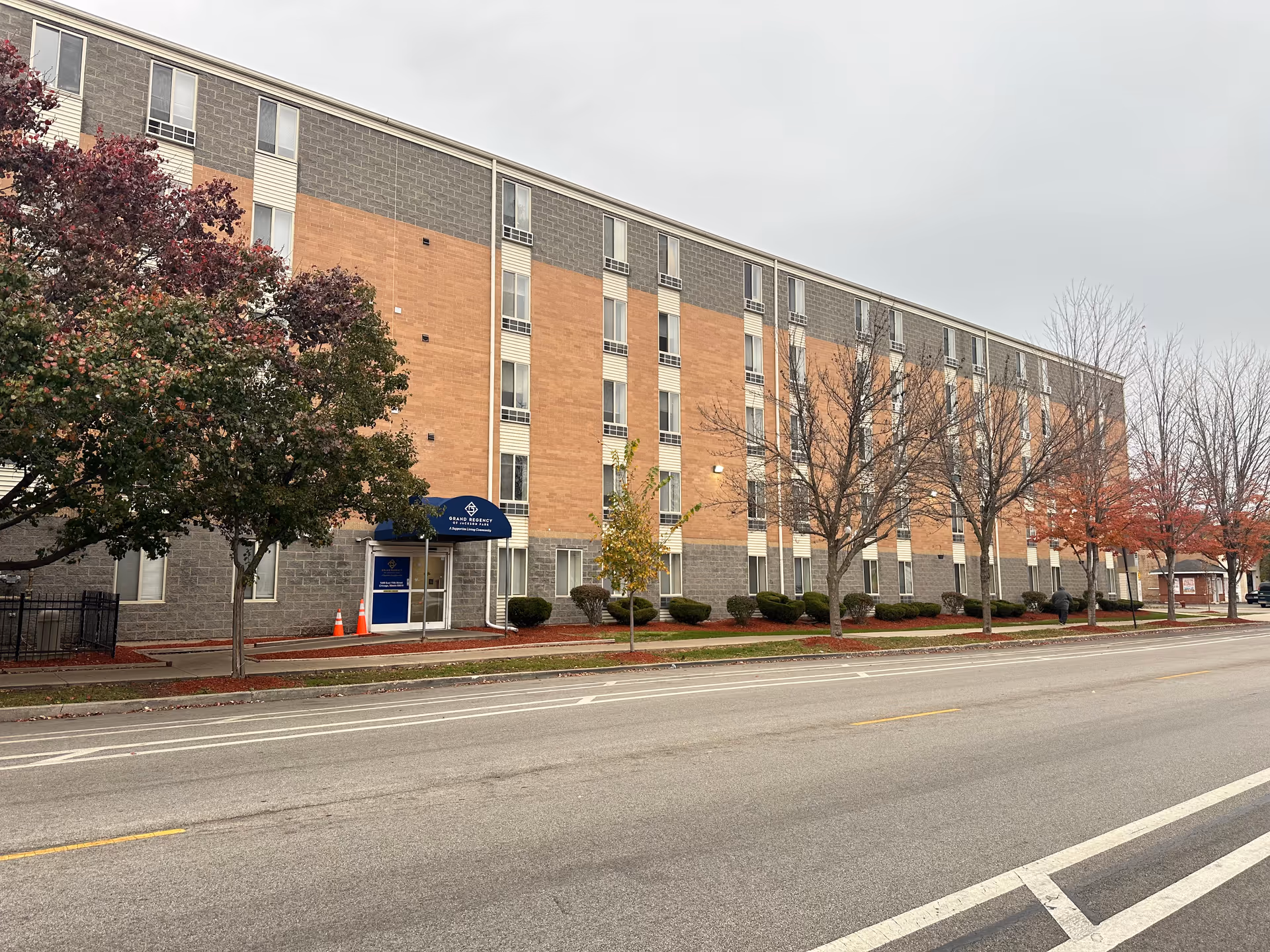 Front exterior of a four-story brick and gray concrete senior living building with a blue awning entrance, trees, and a street in the foreground.