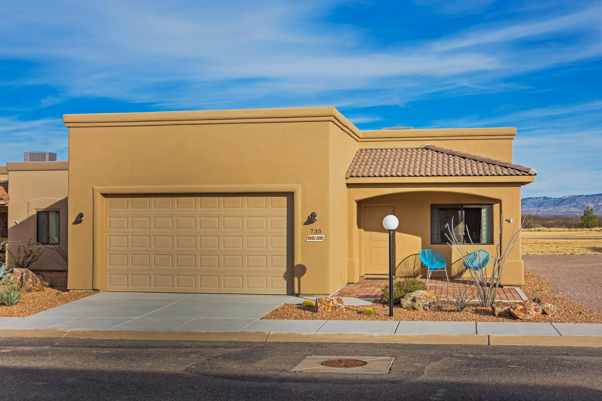 Exterior view of a single-story beige stucco house with a two-car garage, a small covered porch with two turquoise chairs, desert landscaping with rocks and cacti, and a clear blue sky in the background.