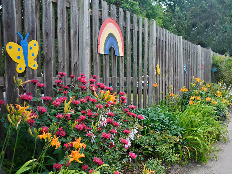 A wooden fence decorated with colorful wooden cutouts including a yellow butterfly, a rainbow, and birds. In front of the fence, there is a garden bed filled with blooming orange and pink flowers and green foliage. Trees and greenery are visible in the background.