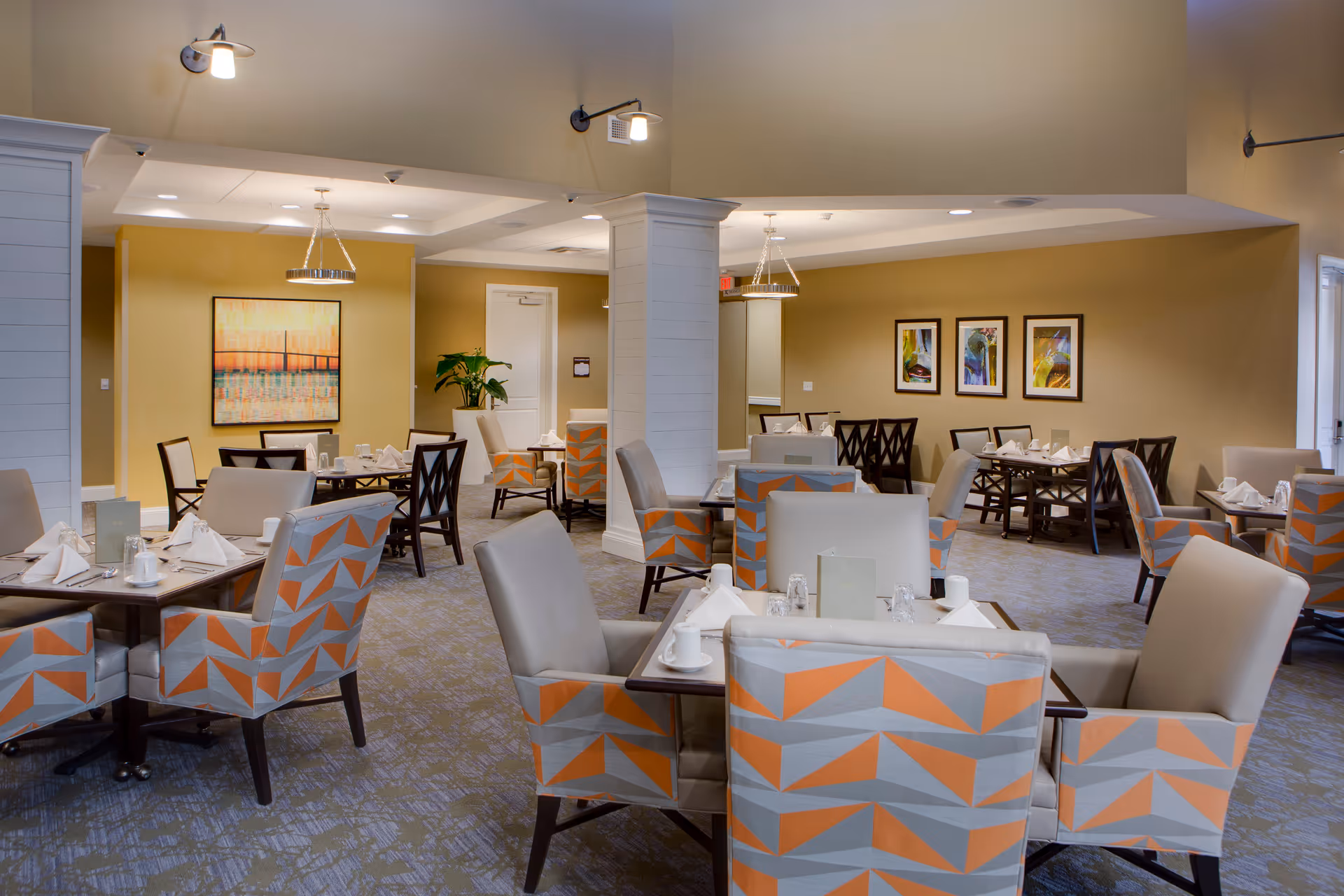 A dining room in a senior living facility with multiple tables set for meals. The chairs have a geometric pattern in orange, gray, and beige. The room has beige walls adorned with framed artwork and is softly lit with ceiling lights and wall sconces. There is a white column in the center and a plant near the back wall.