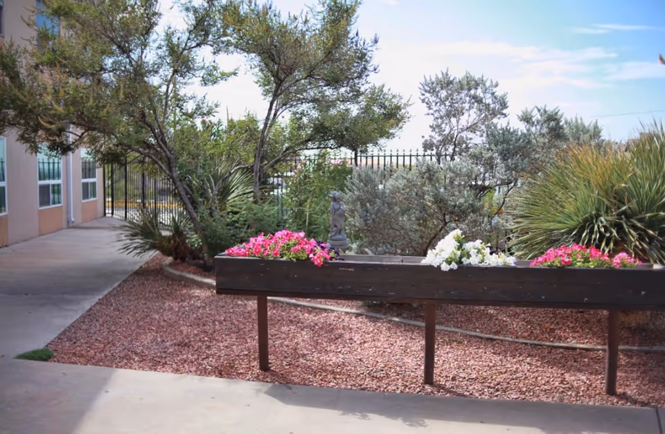 Outdoor garden area at Mountain View Health and Rehabilitation featuring a raised wooden planter box with pink and white flowers, surrounded by various green shrubs and trees. The area is covered with reddish gravel and bordered by a concrete walkway next to a building with windows.
