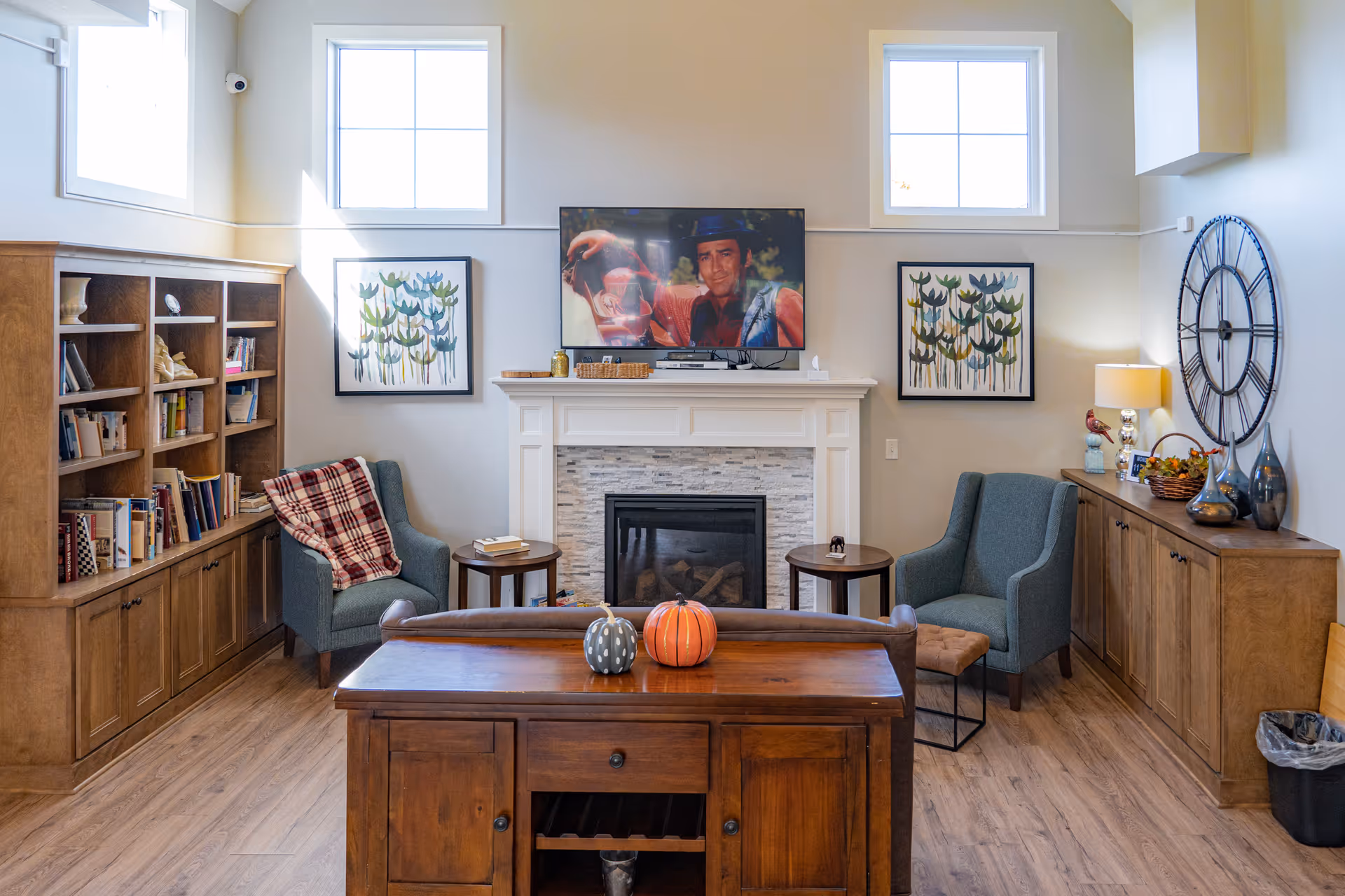 A cozy senior living room with two blue armchairs, a wooden bookshelf filled with books, a wooden cabinet with decorative items, a fireplace with a TV mounted above it, and two framed floral paintings on the wall. The room has wooden flooring and two windows letting in natural light.