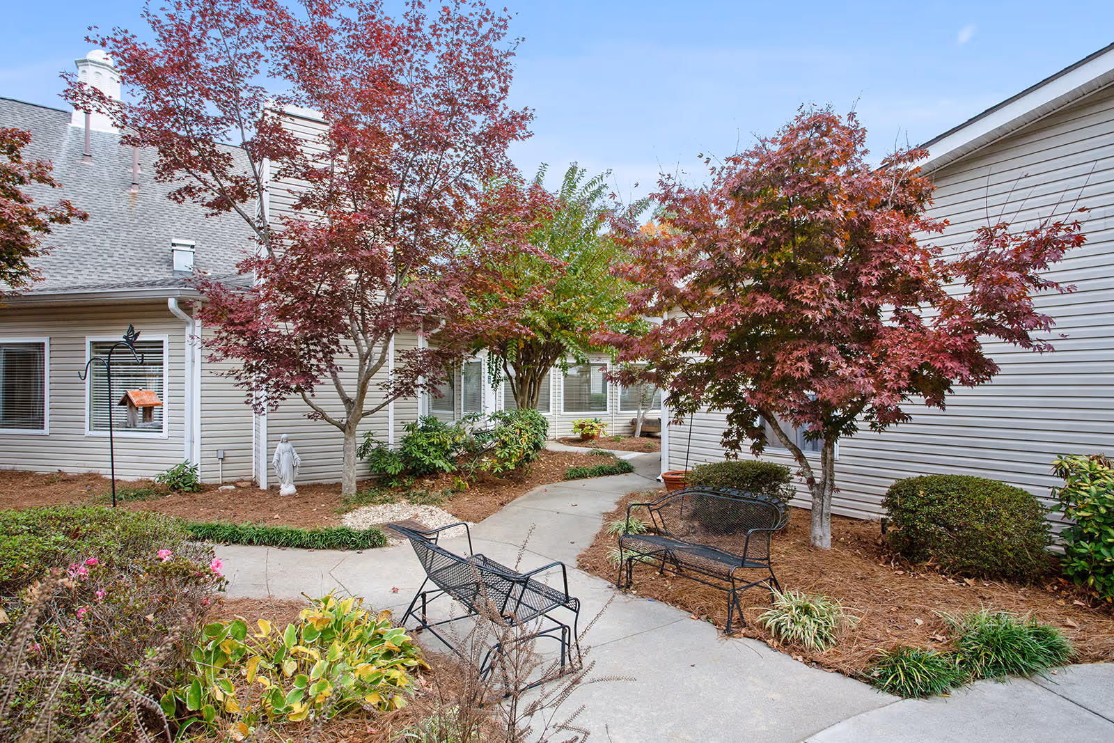 Outdoor courtyard area at Governors Glen featuring a paved walkway, black metal benches, and various trees with red and green foliage surrounded by landscaped bushes and plants, adjacent to beige siding buildings under a clear blue sky.