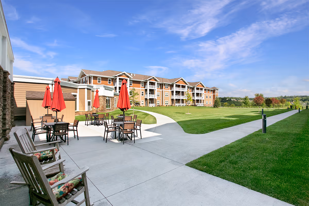 Outdoor patio area with several tables and chairs under red umbrellas, adjacent to a large multi-story residential building with balconies. The area is surrounded by well-maintained green lawns and a concrete walkway under a blue sky with some clouds.