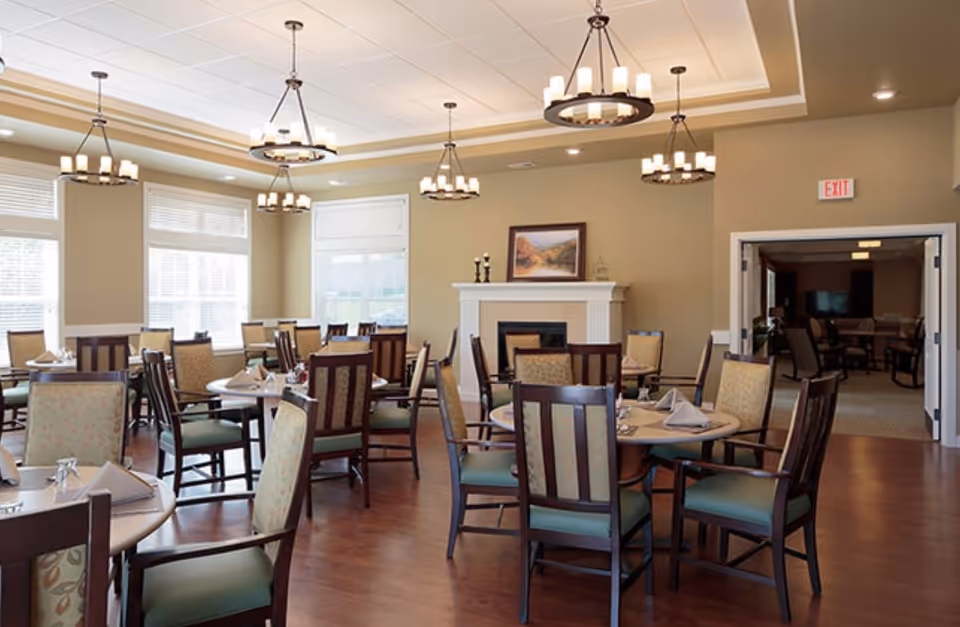 Bright dining room with round tables and wooden chairs, chandeliers, large windows, and a fireplace.